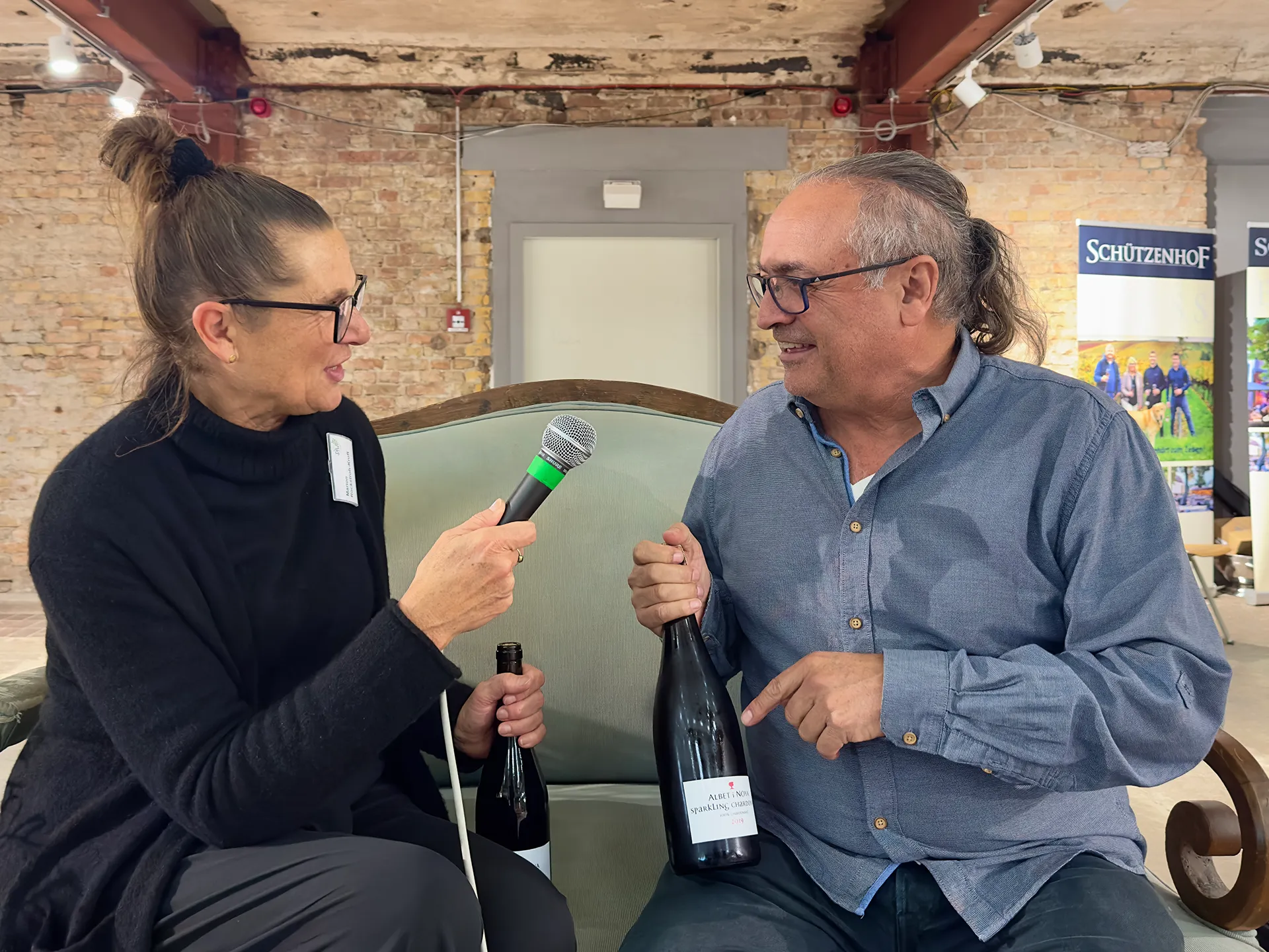 Eine Frau und ein Mann sitzen auf einem Sofa die Frau hält ein Mikrofon der Mann eine Weinflasche in der Hand