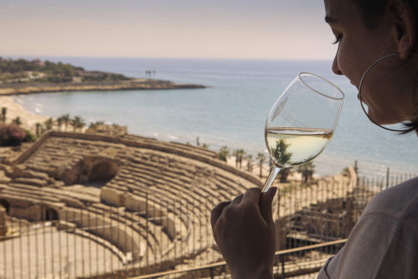 eine Frau hält ein Glas Wein, im Hintergrund das römische Amphitheater von Tarragona